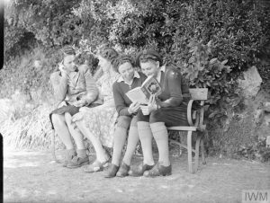 Enjoying a quiet spot on a bench under the trees in the garden of the rest break house in Torquay are (left to right) Mrs Eivemark (the forewoman of a WLA hostel), Miss Mary Pakes, Miss Ann Royne (WLA gang work forewoman) and Mrs Joan Hart (timber corps crane driver). Mrs Eivemark and Miss Pakes are chatting whilst Miss Royne and Miss Hart are reading a book together. Copyright: © IWM. Original Source: http://www.iwm.org.uk/collections/item/object/205200981